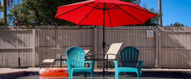 Two Adirondack chairs beside a private pool under a red umbrella, surrounded by green trees on a sunny day